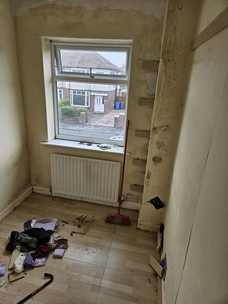 A small bedroom with marked, stained walls and shelf battens screwed to the plaster